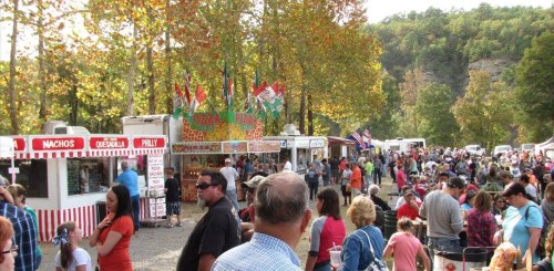 A bustling outdoor festival with food stalls, colorful flags, and crowds enjoying the festivities among autumn trees.