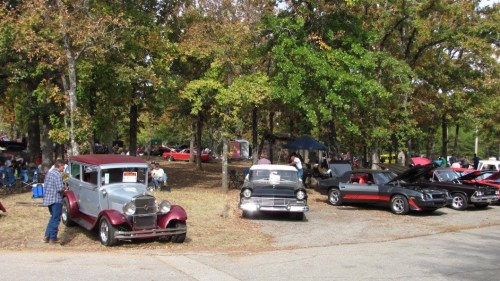 A car show in a park featuring vintage and classic cars, with people mingling among the trees.