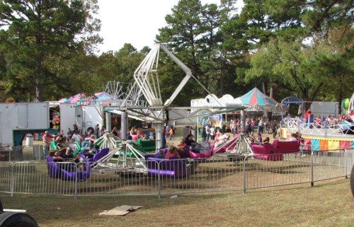 A lively carnival scene with a spinning ride, colorful tents, and people enjoying various attractions in a wooded area.