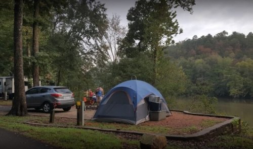 A blue tent set up by a river, with a car nearby and people gathered around a picnic table in a wooded area.