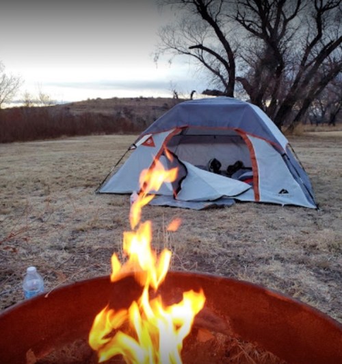 A camping scene with a tent set up near a fire pit, surrounded by open grassland and trees in the background.