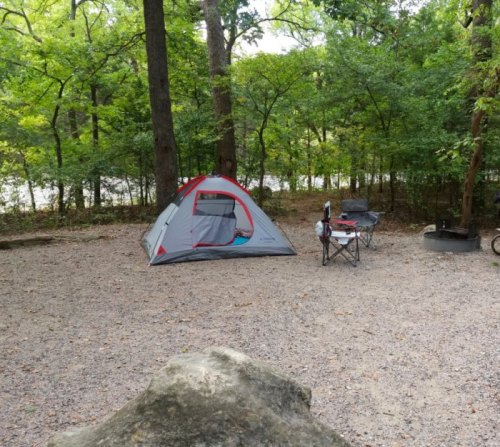 A gray tent set up in a wooded campsite with a chair nearby and a fire pit in the background.