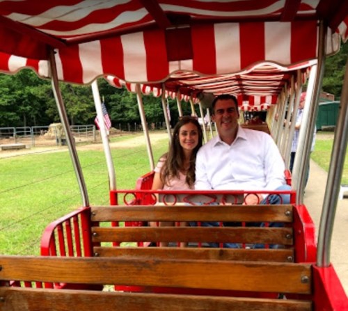 A couple sits together on a red-striped train in a park, surrounded by greenery and a rustic setting.