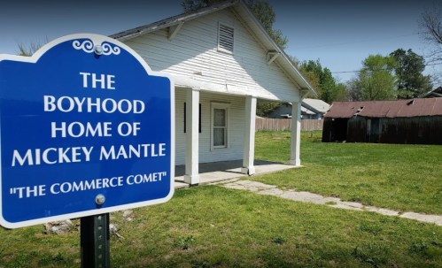 Sign for the boyhood home of Mickey Mantle, with a white house and a weathered shed in the background. Green lawn visible.