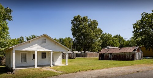 A white house with a porch stands next to a weathered shed, surrounded by green grass and trees.