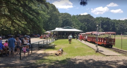 A train station with a red train, surrounded by trees and people waiting in a sunny park setting.