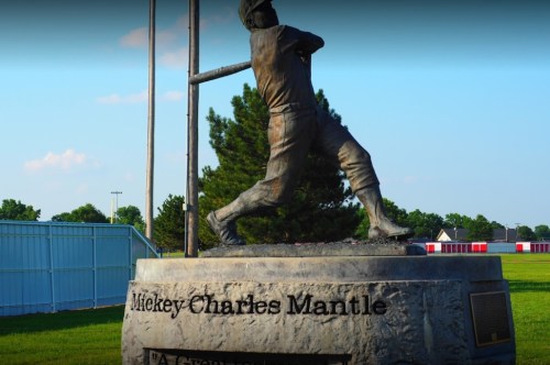 Statue of Mickey Mantle swinging a bat, set in a grassy field with trees and a blue sky in the background.