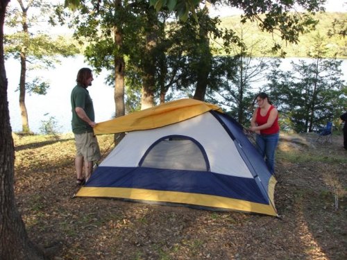 A man and woman set up a blue and yellow tent by a lake, surrounded by trees and nature.