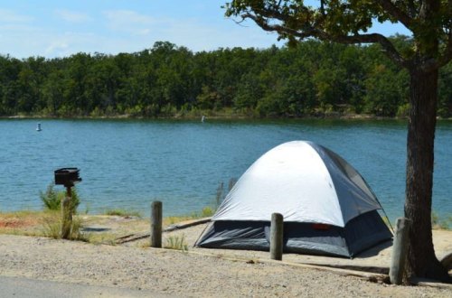 A tent by a calm lake, surrounded by trees, with a grill nearby and a clear blue sky above.