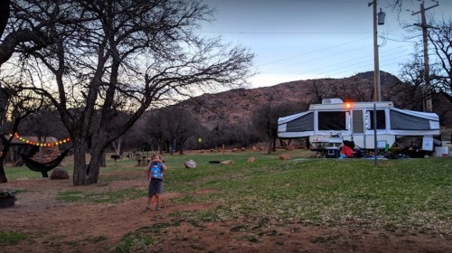 A person walks near a camper in a grassy area surrounded by trees and a mountain in the background during twilight.