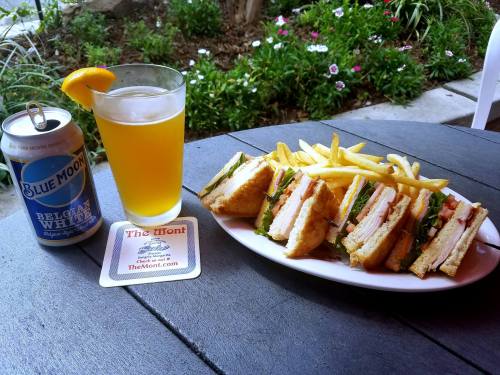 A plate of club sandwiches with fries, a glass of beer, and a coaster on a table surrounded by greenery.