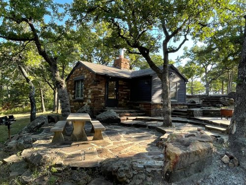 A rustic stone cabin surrounded by trees, featuring a stone patio and picnic table in a serene outdoor setting.