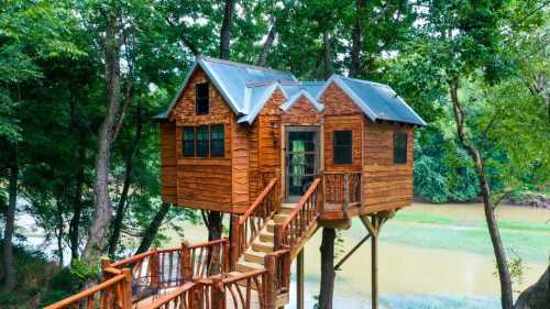 A wooden treehouse with a sloped roof, perched among trees, connected by a wooden walkway over a river.