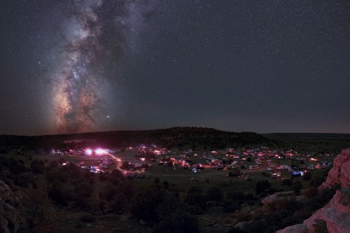 A panoramic view of a starry night sky with the Milky Way above a lit-up campsite in a remote landscape.