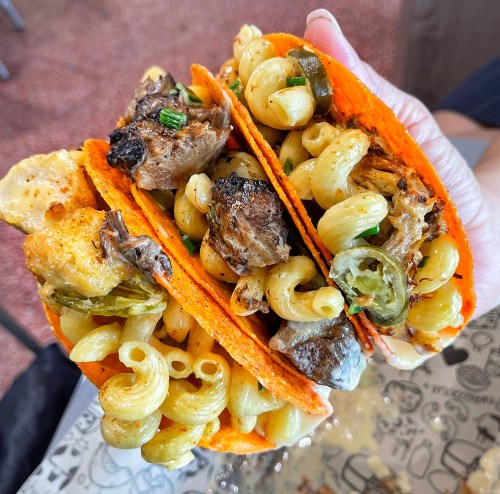 A hand holding three tacos filled with pasta, meat, and jalapeños, all in vibrant orange taco shells.