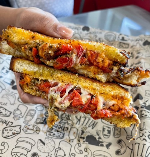 A hand holding a cheesy sandwich filled with roasted red peppers and herbs, served on patterned paper.