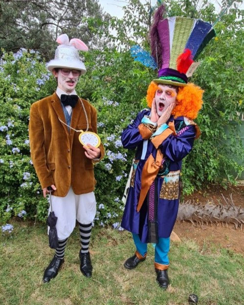 Two whimsical characters in colorful costumes stand in front of greenery, one holding a clock and the other with a large hat.
