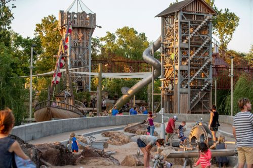 A vibrant playground featuring a pirate ship, tall slide, and children playing on rocks and water features.