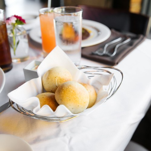 A basket of freshly baked rolls on a dining table, accompanied by drinks and a flower vase.