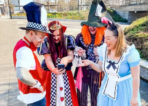 Four people in costumes, including a queen, a mad hatter, and Alice, looking at their phones outdoors.