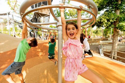 A joyful girl in a pink outfit hangs from a playground structure, while two other children play in the background.