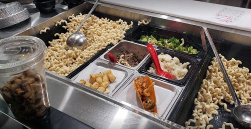 A food counter with curly noodles, various toppings, and containers of meat and vegetables, alongside a cup of snacks.