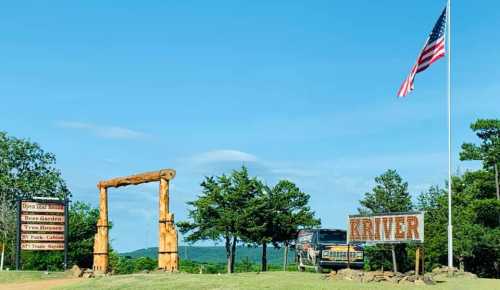 A wooden entrance archway with signs, a truck, and an American flag against a clear blue sky and green trees.