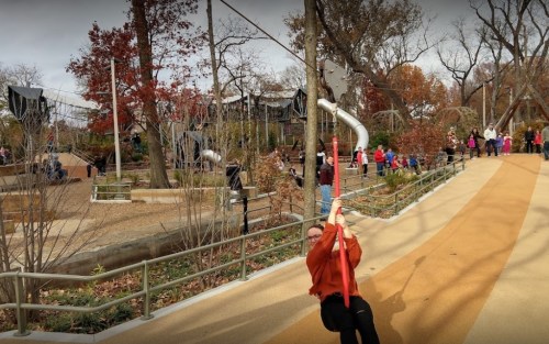 A child on a red zip line in a park, surrounded by trees and people enjoying the playground.