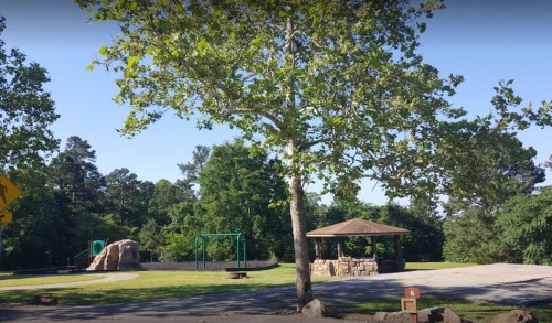 A park scene featuring a large tree, playground equipment, a stone gazebo, and green grass surrounded by trees.