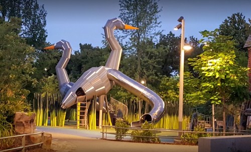 A unique playground structure resembling two large, metallic birds with slides, surrounded by greenery and evening lights.