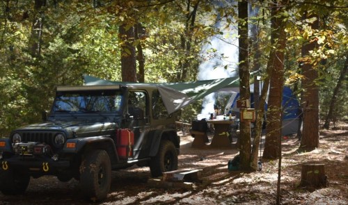 A green Jeep parked near a campsite with a tent and smoke rising from a fire in a wooded area.
