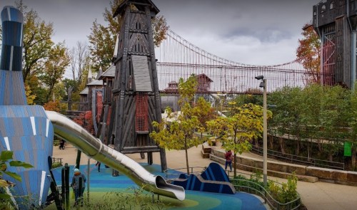 A colorful playground with a slide, surrounded by trees and structures, with a bridge in the background under a cloudy sky.