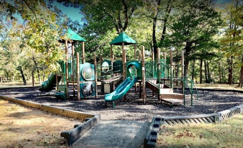 A playground with green slides and structures surrounded by trees and gravel, set in a sunny outdoor area.