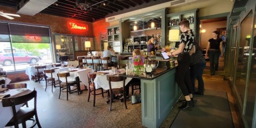 A cozy restaurant interior with tables, a bar area, and patrons dining and working. A neon sign adds ambiance.