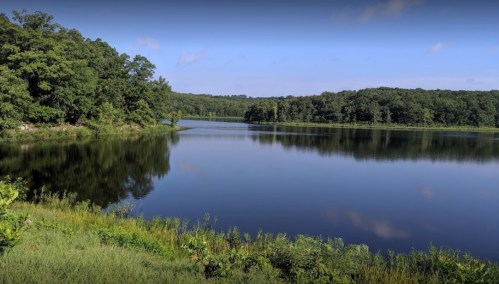 A serene lake surrounded by lush greenery under a clear blue sky, reflecting the landscape in calm waters.