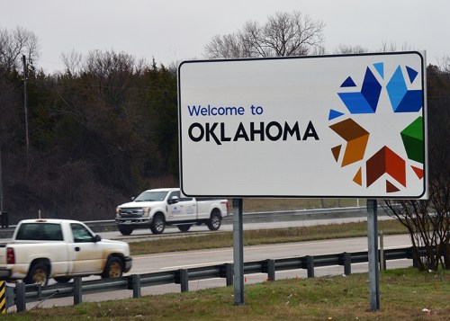 A roadside sign welcoming travelers to Oklahoma, featuring a colorful star design and a passing white truck.