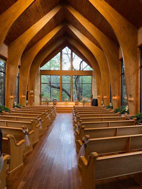 Interior of a wooden chapel with high arched ceilings, rows of pews, and large windows overlooking trees.