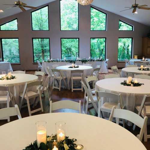 A spacious event room with large windows, round tables set with white linens, and greenery centerpieces.