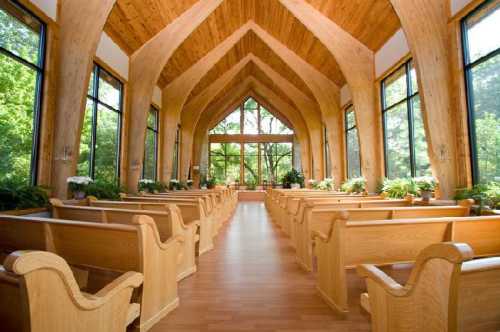 Interior of a modern chapel with wooden beams, rows of pews, and large windows overlooking greenery.