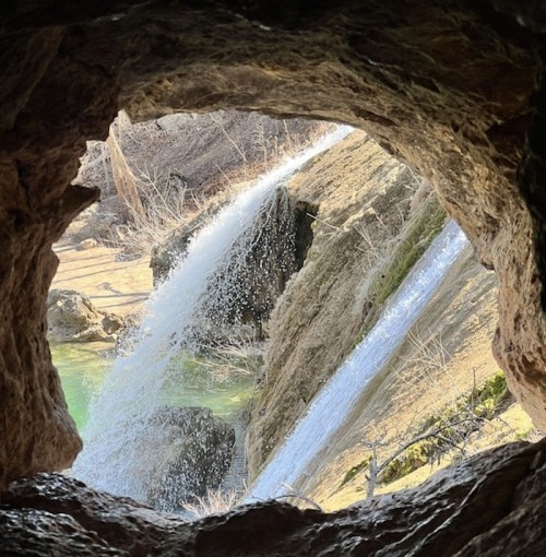View of a waterfall cascading through a rocky opening, surrounded by greenery and natural terrain.