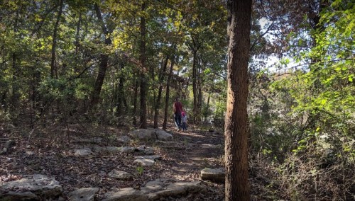 A person walks along a wooded trail with a child, surrounded by trees and rocky terrain.