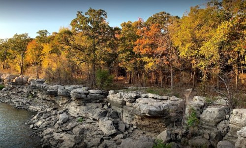 Rocky shoreline with autumn trees in vibrant colors along a calm body of water.