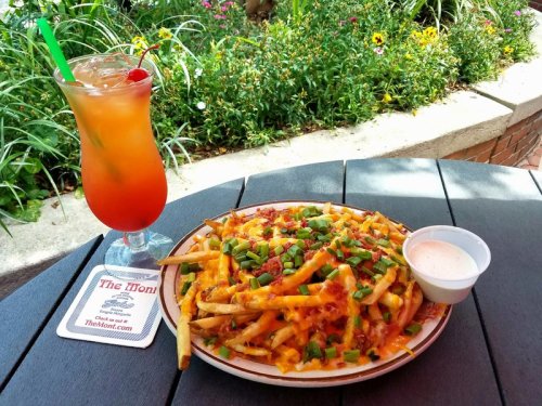A plate of loaded fries topped with cheese and green onions, alongside a colorful cocktail on a table with flowers in the background.