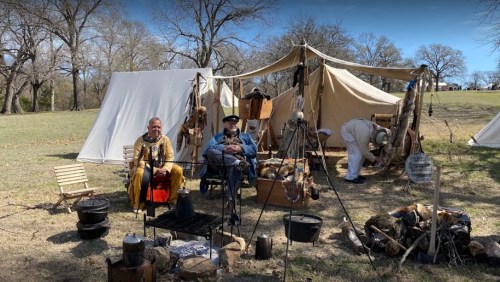 Two men in historical attire sit by a tent, with a campfire setup and a third person preparing nearby.