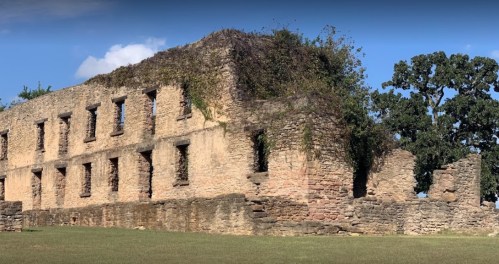 Ruins of a stone building covered in vines, set against a clear blue sky and green trees.