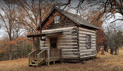 A rustic log cabin with a wooden porch, surrounded by autumn trees and fallen leaves.