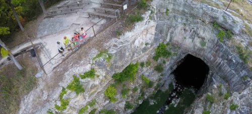 Aerial view of a deep, rocky pit with a dark opening, surrounded by trees and a viewing area with people.