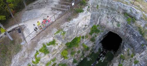 Aerial view of a deep, rocky pit with a dark opening, surrounded by trees and a viewing area with people.