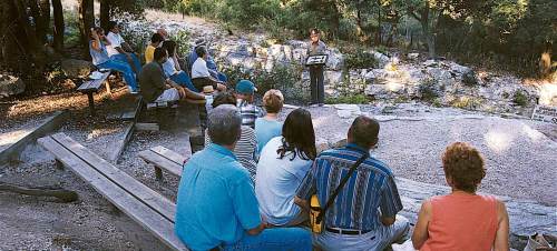 A group of people seated outdoors, listening to a speaker at a podium in a natural setting.
