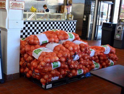 A large stack of bags filled with onions sits on a pallet in a grocery store, with a counter and a worker in the background.